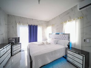 A modern bedroom with a marble-patterned wall, a tufted headboard, a flat-screen TV, and a bedside table with drawers. - 8BR Private Villa in Sosúa - 2 Mins From Nightlife (Sosúa)