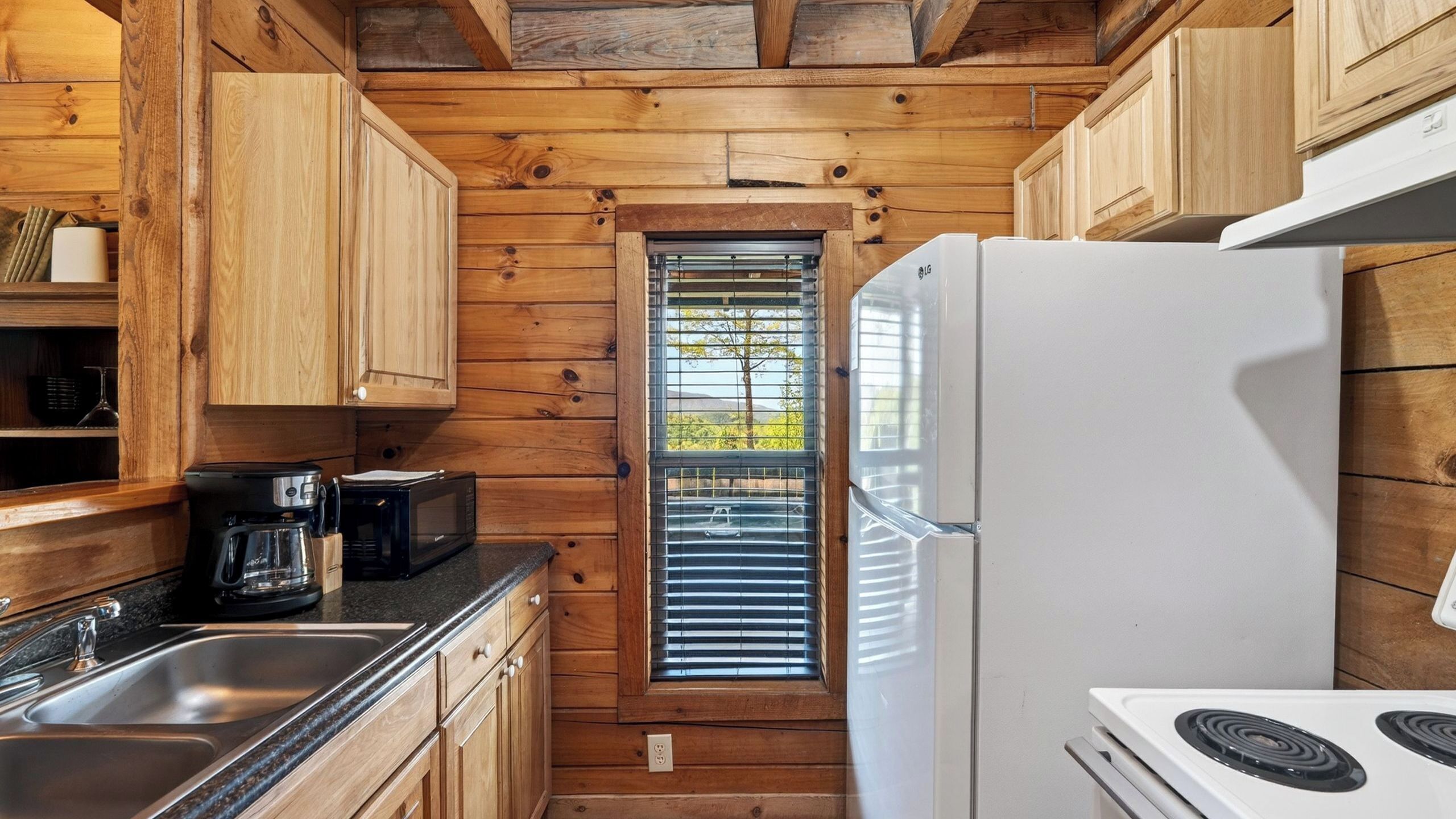 A compact kitchen with wooden cabinetry, a white refrigerator, a stove, and a window with blinds.