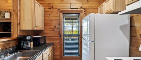 A compact kitchen with wooden cabinetry, a white refrigerator, a stove, and a window with blinds.