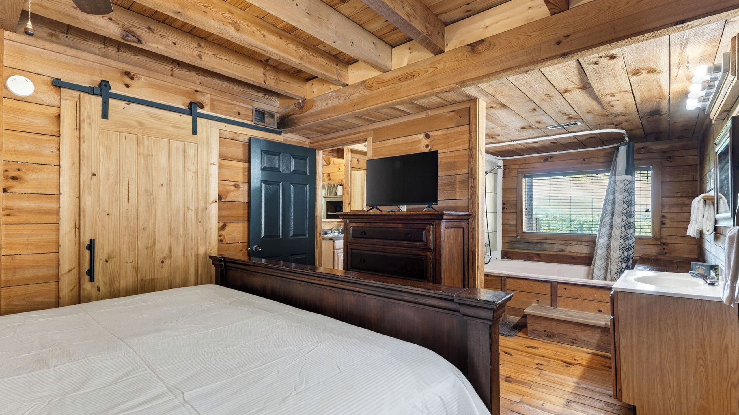 A wooden cabin bedroom with a bed, a TV, a small kitchenette, and a bathroom with a window.