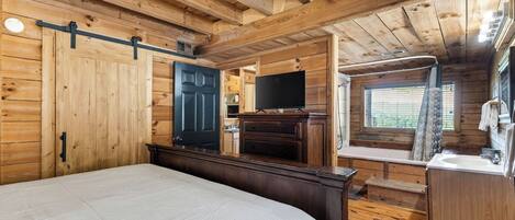 A wooden cabin bedroom with a bed, a TV, a small kitchenette, and a bathroom with a window.