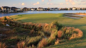 Ein Golfplatz mit einem Wassengraben, Häusern im Hintergrund und einer Fahne auf dem Grün.