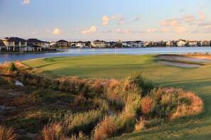 Ein Golfplatz mit einem Wassengraben, Häusern im Hintergrund und einer Fahne auf dem Grün.