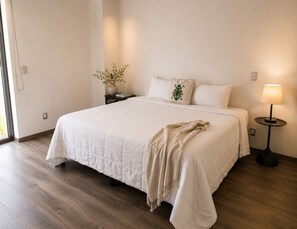 A neatly made bed with a white duvet, a bedside table with a lamp, and a vase with a plant on the nightstand. - 3BR · Country Club Guadalajara · cocina · rooftop bar · WiFi (guadalajara)