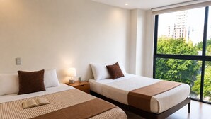 A hotel room with two beds, a nightstand with a lamp, and a large window offering a view of buildings and trees. - Cama King · rooftop bar · Country Club Guadalajara · limpieza diaria (guadalajara)