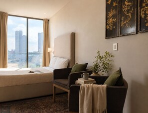 A hotel room with a bed, a sofa, a chair, a coffee table, a vase with plants, and a city view. - Suite Premier · vistas · Country Club Guadalajara · limpieza diaria (guadalajara)