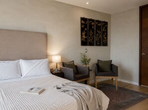 Een slaapkamer met een bed, nachtkastje, lamp, twee fauteuils, een decoratief paneel en een deur.