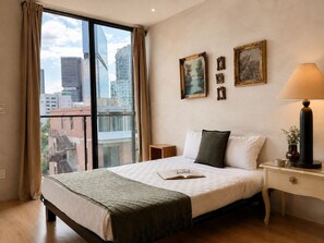 A hotel room with a large window, a bed with a green throw, a nightstand with a lamp, and framed pictures on the wall. - Suite Premier Grand · vistas · Country Club GDL · limpieza diaria (guadalajara)