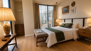 Ein Hotelzimmer mit Bett, Nachttischen, einer Bank, einem Fenster mit Vorhängen, einem Deckenventilator und Kunstwerken an der Wand.