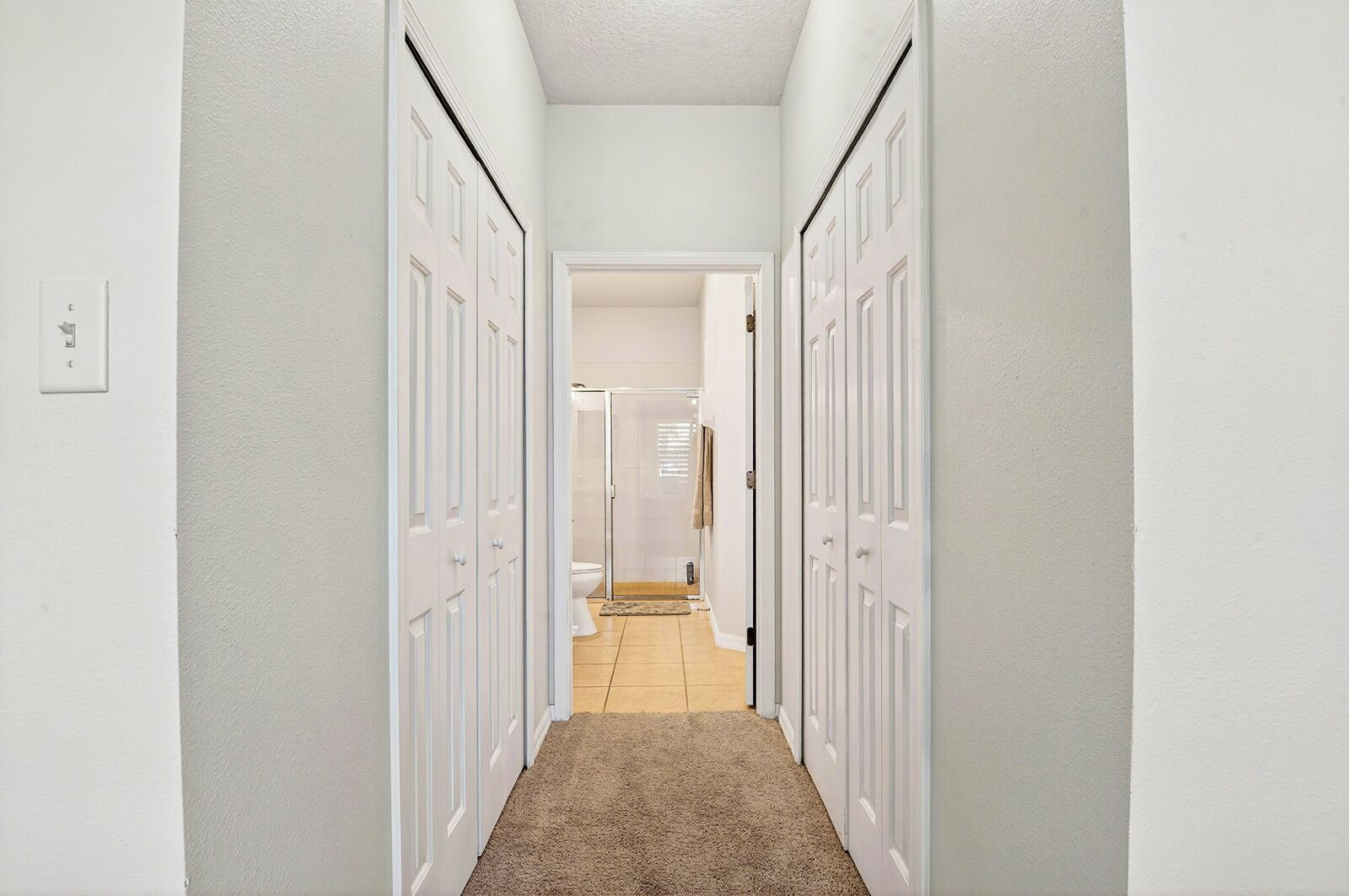 A hallway with white walls, a carpeted floor, and white doors leading to a bathroom and another room.