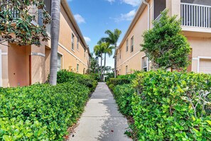 A narrow pathway flanked by lush green hedges and multi-story buildings with balconies. - Fantastic location , 3BD/2BT Condo with garage,patio and pool in Gated community (Sarasota)