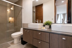 A modern bathroom with a glass shower enclosure, a toilet, a vanity with two sinks, and a mirror above the vanity. - Estudio Premier · vistas · Country Club GDL · limpieza diaria (guadalajara)