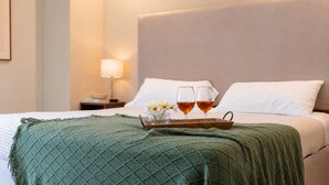 Een net opgemaakt bed met een groene deken, twee glazen amberkleurige vloeistof en een dienblad met bloemen in een hotelkamer.