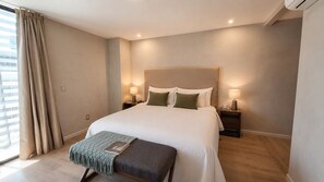 A bedroom with a bed, bedside tables, a bench, and a window with curtains. - Estudio Premier · vistas · Country Club GDL · limpieza diaria (guadalajara)