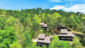 A forested area with multiple cabins, each with a distinct roof design, surrounded by dense green trees. - Anna's Cabin by Avantstay Private Hot Tub + Mountain Views + Covered Deck 7 Min to Townsend (Townsend)