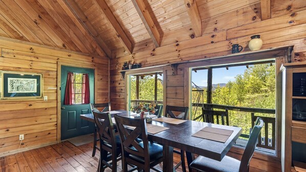 A wooden cabin interior with a dining table, chairs, a green door, and a view of greenery outside.