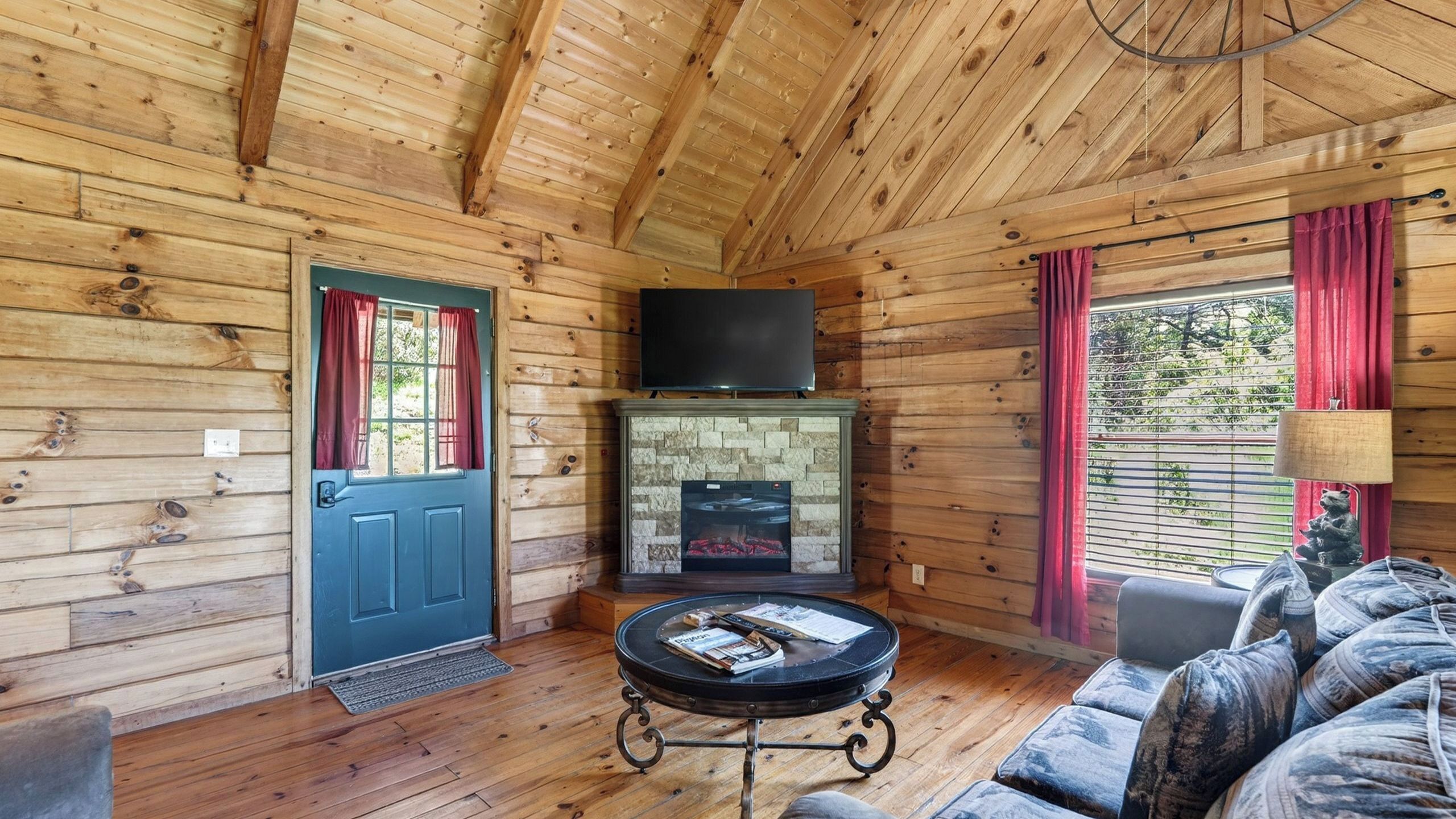 Interior de una cabaña de troncos con chimenea, un televisor montado encima, una mesa redonda de centro con libros y un sofá con cojines estampados.