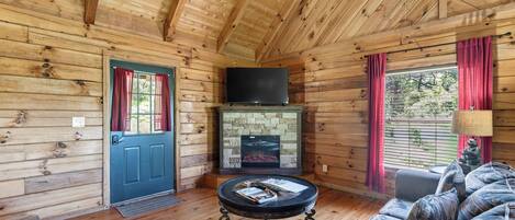 Interior de una cabaña de troncos con chimenea, un televisor montado encima, una mesa redonda de centro con libros y un sofá con cojines estampados.