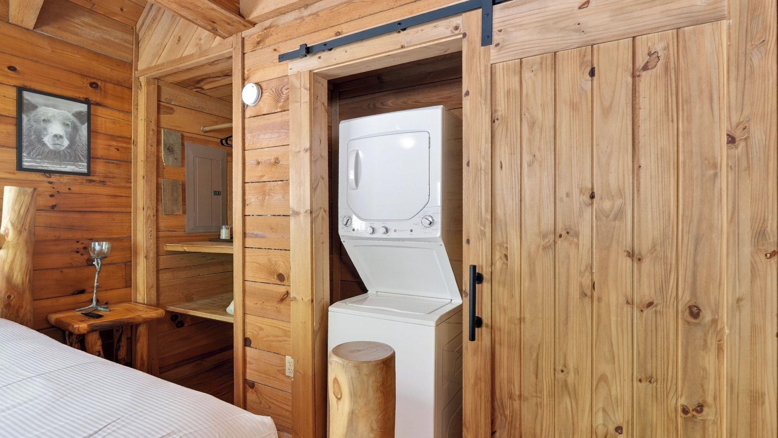 A wooden cabin interior with a bed, a small table with a metal cup, and a white washing machine.