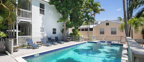 A swimming pool with lounge chairs, a wooden deck, and a white building with a balcony.