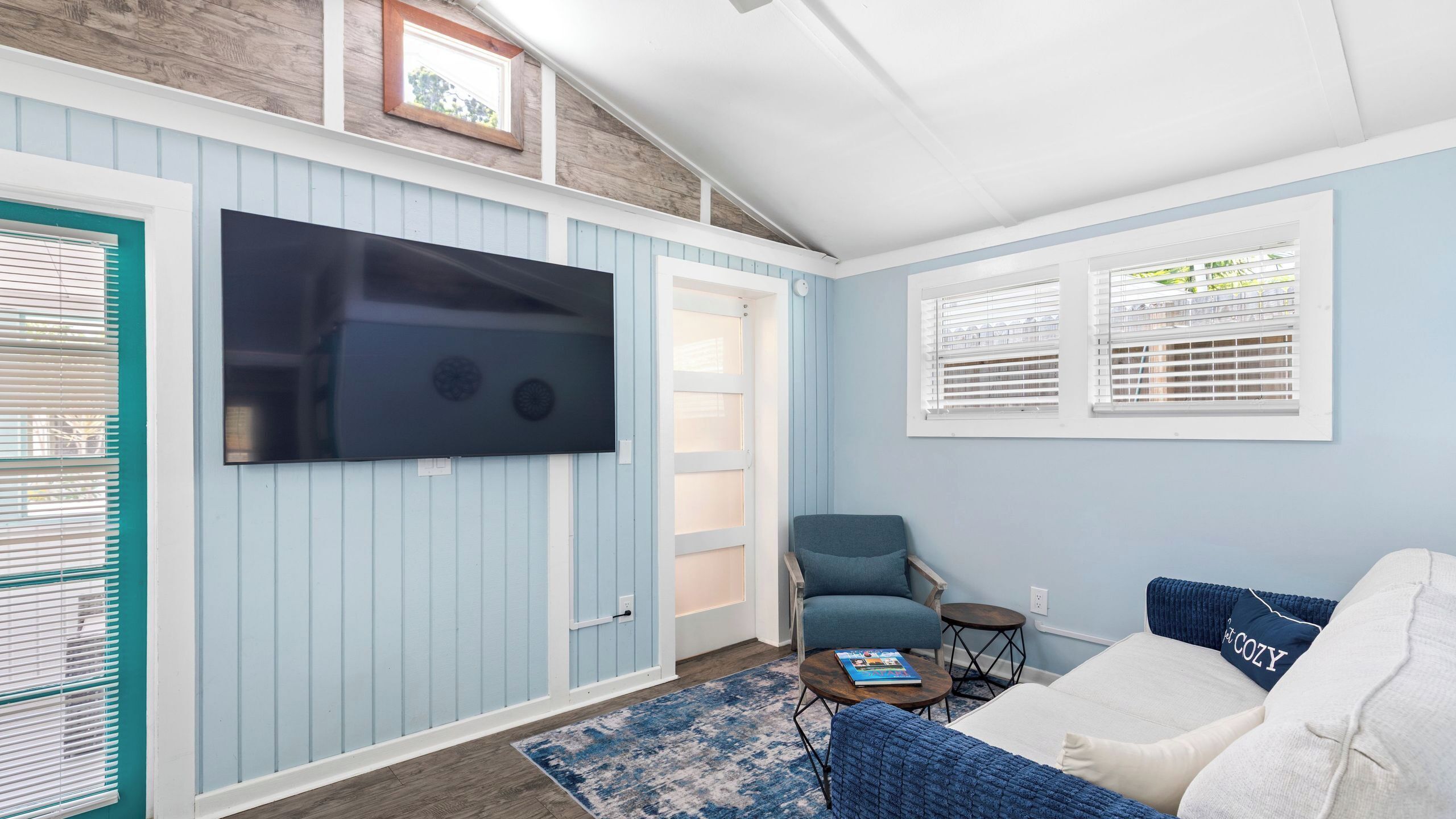 Habitación con una televisión montada en la pared, un sofá azul, una mesita y una puerta que da a un espacio exterior.
