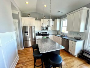 A kitchen with white cabinets, stainless steel appliances, a granite countertop, and a bar stool. - Bee by the Sea, 5 BR house 1 minute to the beach, private road near restaurants (Oak Island)