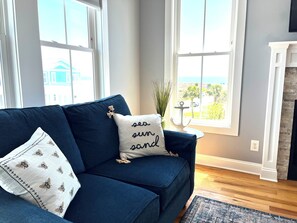 A living room with a dark blue sofa, a white pillow with a bee pattern, and a cushion with 'sea sun sand' text. - Bee by the Sea, 5 BR house 1 minute to the beach, private road near restaurants (Oak Island)