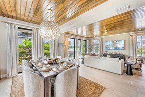 A modern living room with a dining area, wooden ceiling, large windows, and a flat-screen TV.