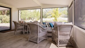 Ein Zimmer mit Korbmöbeln, inklusive Tisch und Stühlen, großen Fenstern und Blick auf Grünfläche.