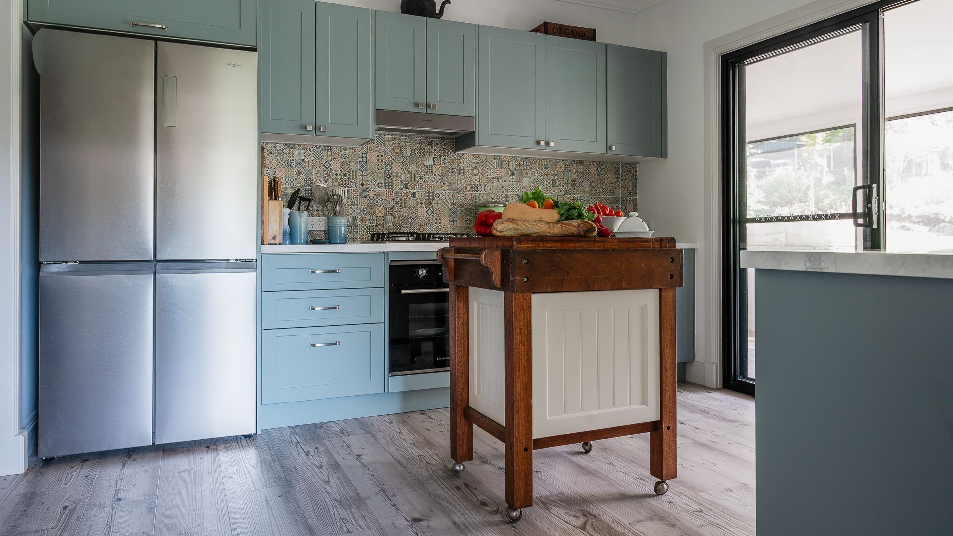 A kitchen with teal cabinets, a stainless steel refrigerator, an oven, and a wooden cart with produce.
