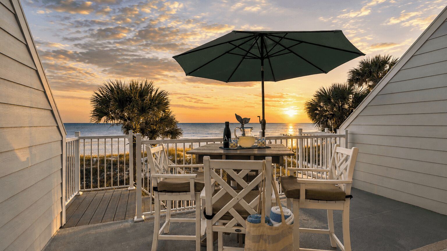 Eine Terrasse mit Tisch, Stühlen, einem Sonnenschirm und Blick auf den Sonnenuntergang über dem Meer.