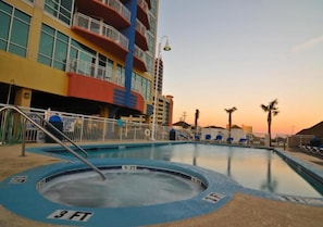 Una piscina con una garita de salvavidas, un edificio de varios pisos con balcones y palmeras al fondo.