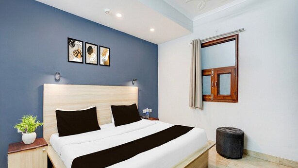 A modern hotel room with a large bed, a bedside table with a plant, a small ottoman, and a window with curtains. - Super Collection O Sector 20 Noida (Noida)
