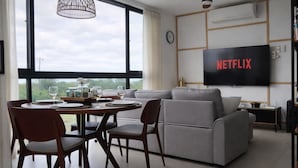 Un salon moderne comprenant un coin repas, un canapé gris, un téléviseur fixé au mur et une grande fenêtre offrant une vue dégagée.