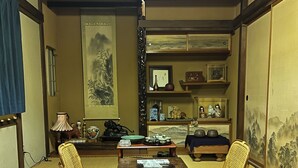 Um quarto japonês tradicional com tatames, uma mesa baixa, duas cadeiras, um candeeiro pendente e uma parede com um quadro emoldurado.