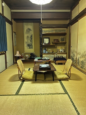 Une piĂšce de style japonais traditionnel, avec un sol recouvert de tatamis, une table basse, deux chaises, un luminaire suspendu et un mur ornĂ© dâun tableau encadrĂ©.