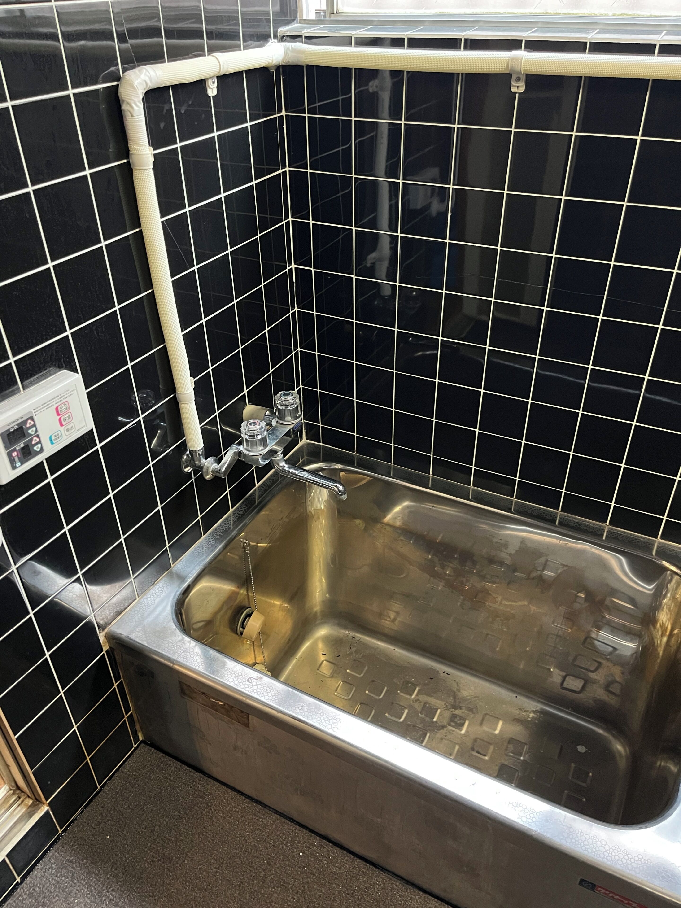Une salle de bain avec des murs recouverts de carreaux noirs, un évier en acier inoxydable et un tuyau blanc qui longe le mur.