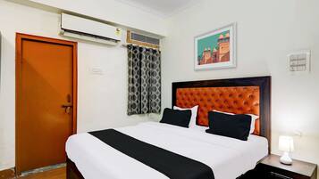 Een hotelkamer met een bed, een airconditioner, een deur, een gordijn, een nachtkastje met een lamp en een wanddecoratie.