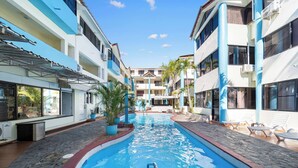 Una piscina con escalera, rodeada de sillas de descanso y plantas en macetas, con edificios a ambos lados.