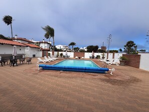Ein Swimmingpool mit Liegestühlen und Tischen, umgeben von einer Ziegelterrasse und weißen Gebäuden mit roten Ziegeldächern.
