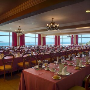 Une salle de banquet avec des tables dressées pour le repas, de grandes fenêtres ornées de rideaux et des lustres.