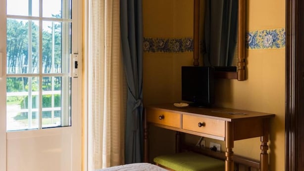 Habitación de hotel con cama, escritorio de madera con televisor, pequeño banco y ventana con cortinas.