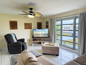 Ein Wohnzimmer mit Deckenventilator, einem Flachbildfernseher, einem grauen Sessel, einem beigen Sofa, einem kleinen Hocker und einem Fenster mit Vorhängen.