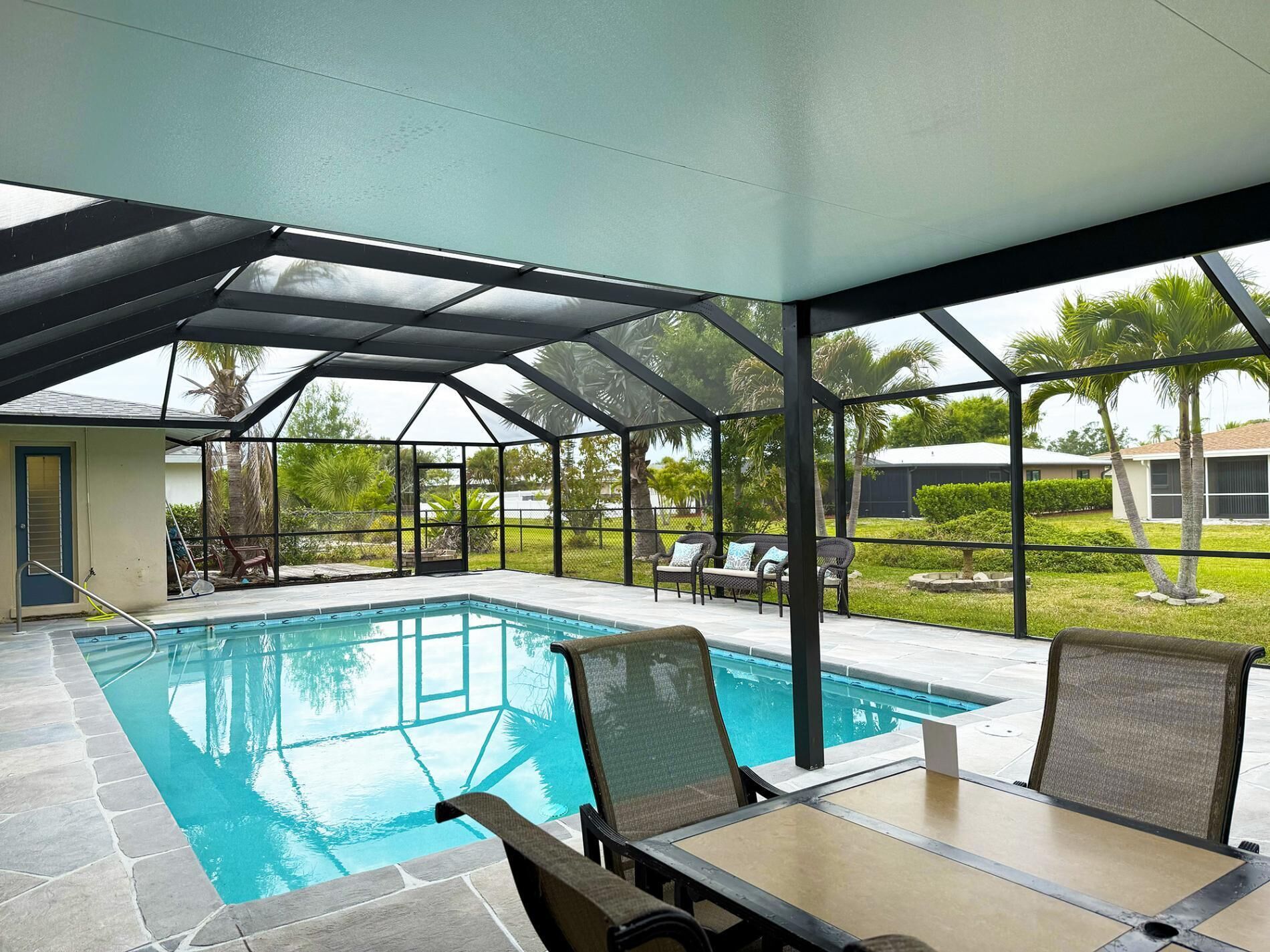 A covered outdoor swimming pool with lounge chairs and a table, surrounded by a fenced area with palm trees.