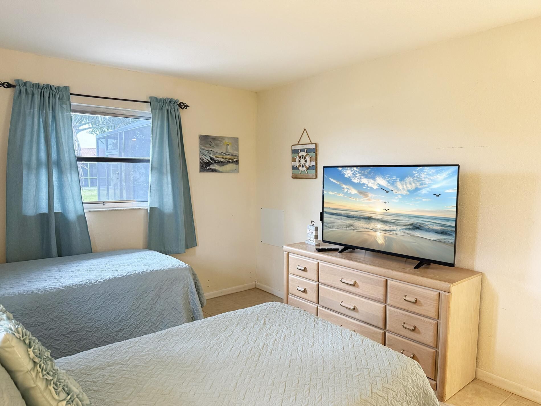 Un dormitorio con dos camas, un televisor sobre un aparador, una ventana con cortinas y un cuadro con temática náutica en la pared.