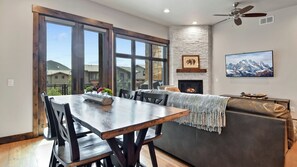A modern living room with a fireplace, a dining area with a large window, and a view of the outside. - Alpine Abode by Avantstay 5min Walk To Lift Base! (Park City)