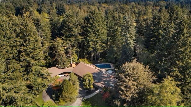 A house surrounded by dense forest, with a visible swimming pool and a driveway leading to the entrance.