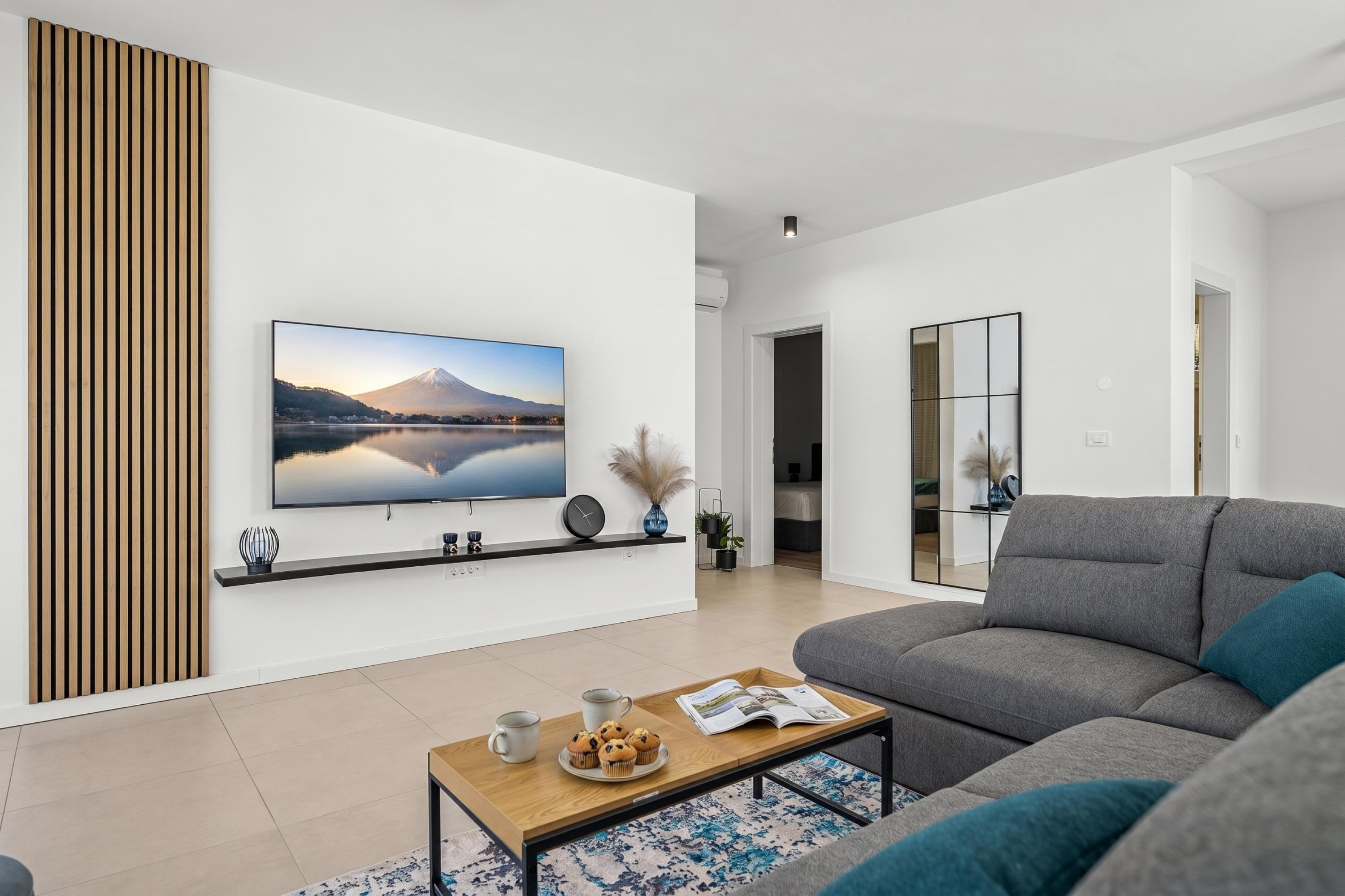 A modern living room with a flat-screen TV, a grey sofa, a coffee table with a tray of muffins, and a wooden slat wall panel.