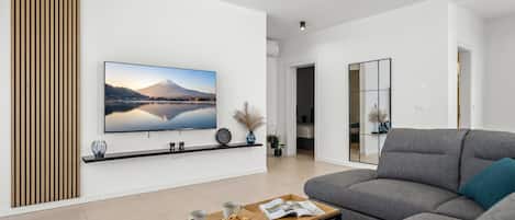 A modern living room with a flat-screen TV, a grey sofa, a coffee table with a tray of muffins, and a wooden slat wall panel.
