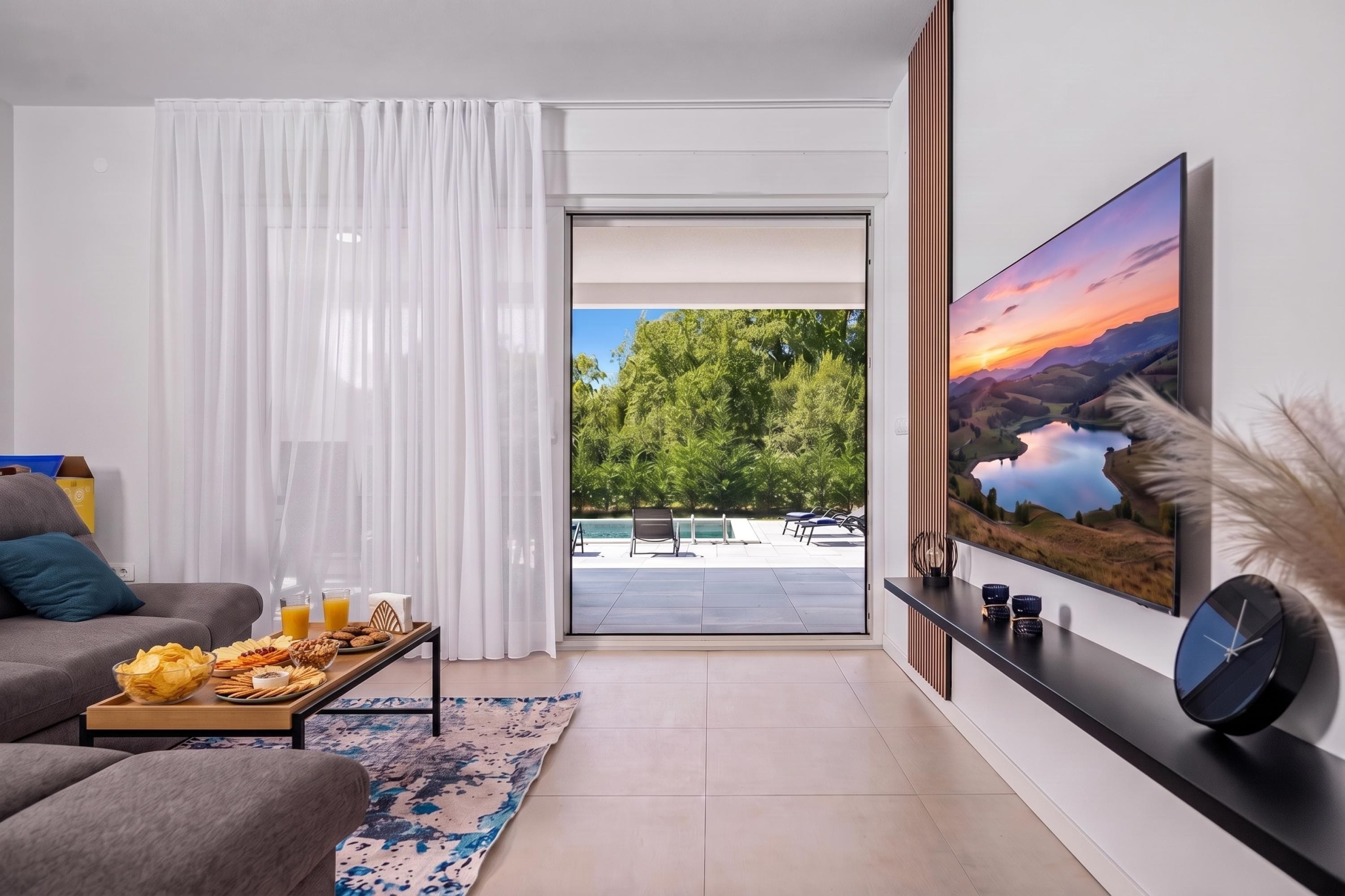 A modern living room with a sofa, a dining table with snacks, a painting, and a view of an outdoor pool area.